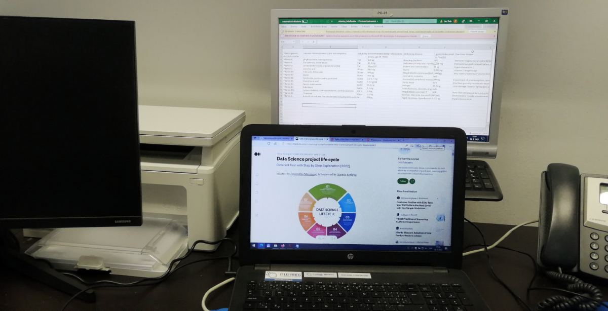 pracovisko s notebookom, tlačiarňou a dvoma počítačmi s použitím Excelu