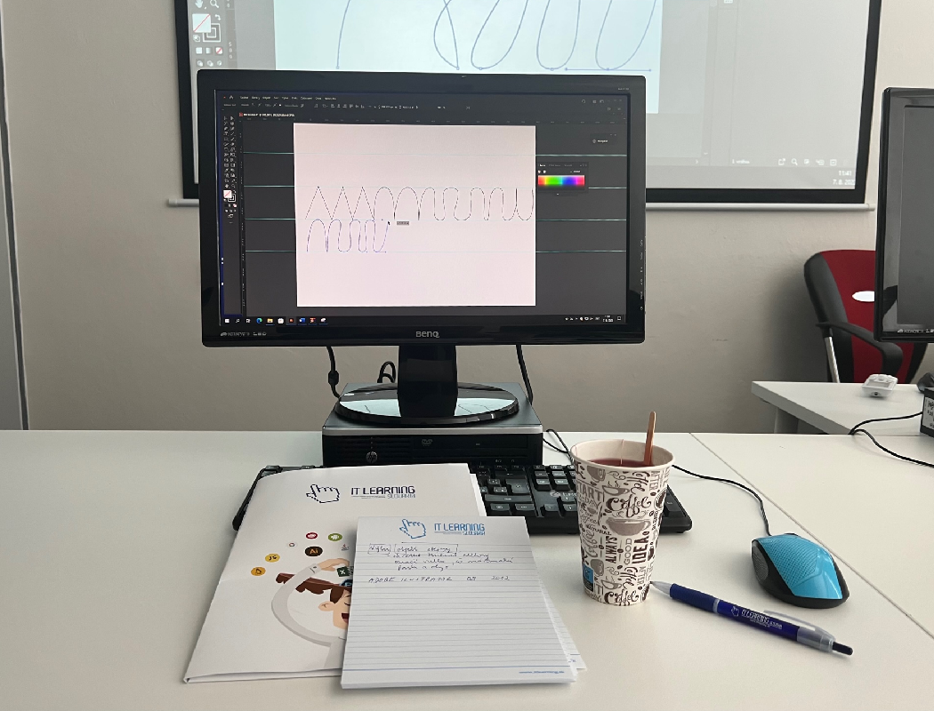 Monitor, vedľa ktorého sú učebné materiály z kurzu Adobe Illustrator 1. Vedľaa monitoru sa nachádza pero, čaj a počítačová myš.